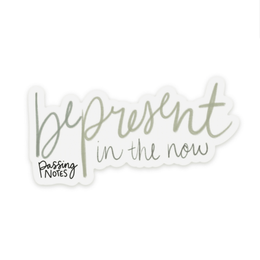 Clear backed inspirational sticker with handwritten green font that says "be present in the now"