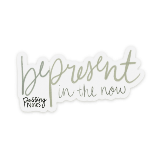 Clear backed inspirational sticker with handwritten green font that says "be present in the now"