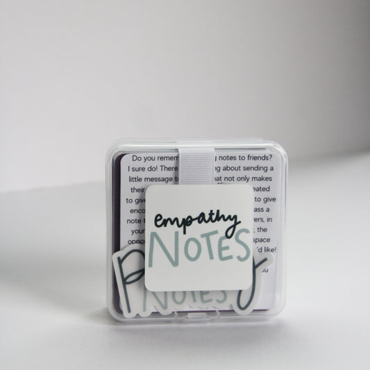 A small clear box labeled "Empathy Notes" rests on a white surface, slightly open to reveal encouraging notes inside. Set against a plain, light-colored wall, this subtle support system invites moments of connection and understanding.