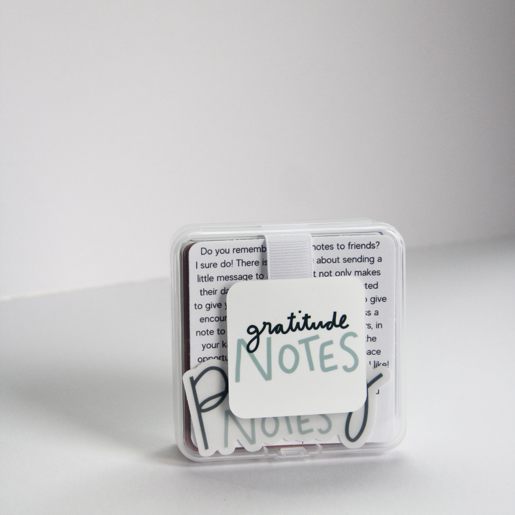 A small, clear box labeled "Gratitude Notes" contains note cards filled with gratitude affirmations. The background is plain white, and a few cards are slightly visible inside the box.