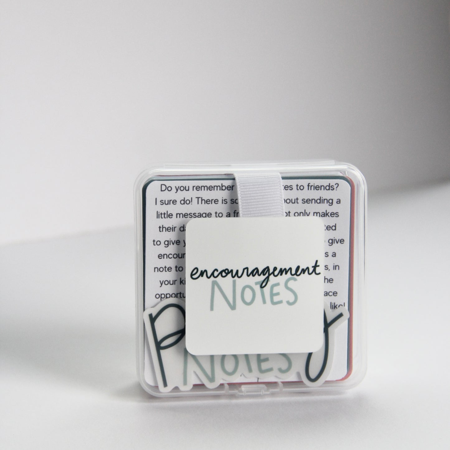 A small transparent box with a white band holds a stack of "Encouragement Notes," perfect for a quick pick-me-up. The minimalist white background accentuates its clean and uplifting design, ready to lift spirits whenever needed.