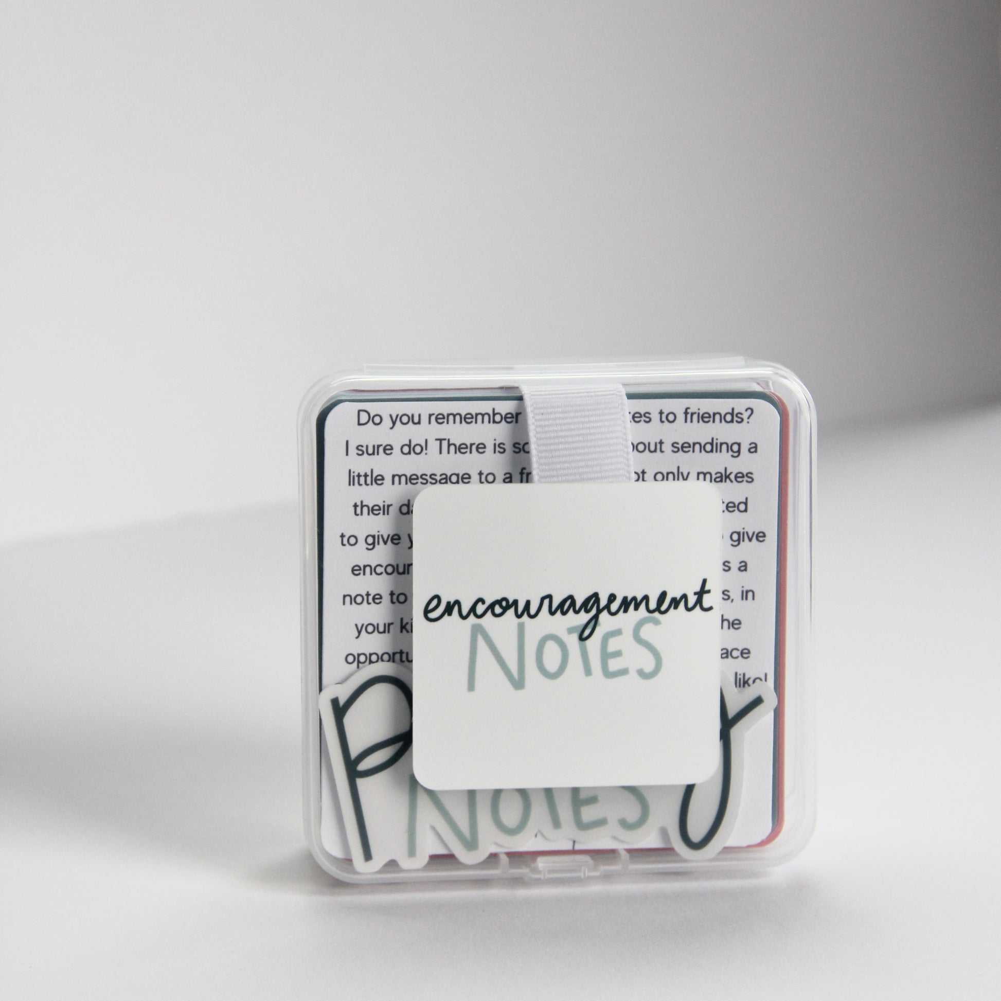 A small transparent box with a white band holds a stack of "Encouragement Notes," perfect for a quick pick-me-up. The minimalist white background accentuates its clean and uplifting design, ready to lift spirits whenever needed.