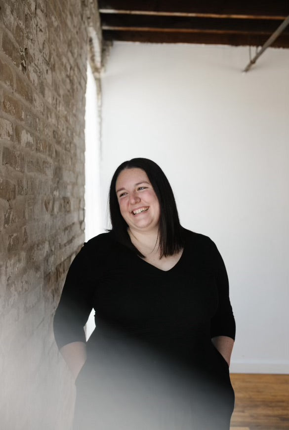 Photo of Bethany, the artist and owner of Passing Notes Studio standing next to a brick wall. She is smiling, looking away from the camera and wearing a black dress.