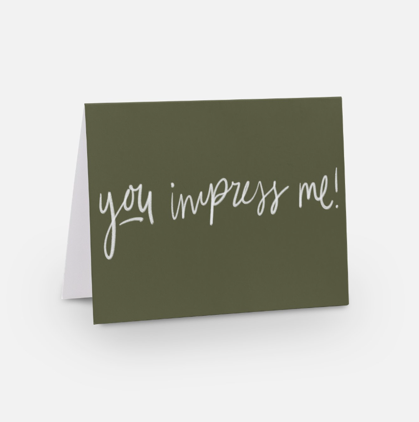 A green encouragement card with white handwritten text that reads, "you impress me!" radiates positivity as it stands upright on a white surface.