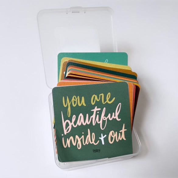 A box of colorful encouraging notes with the top card displaying "you are beautiful inside + out" in white and yellow handwriting on a dark green background. These note cards are neatly arranged inside a small, clear plastic carrying case, making it easy to spread encouragement wherever you are.