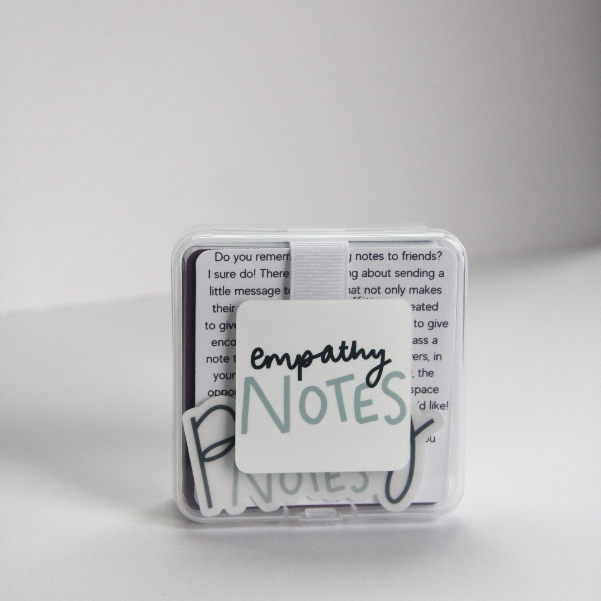 A small clear box labeled "Empathy Notes" rests on a white surface, slightly open to reveal encouraging notes inside. Set against a plain, light-colored wall, this subtle support system invites moments of connection and understanding.