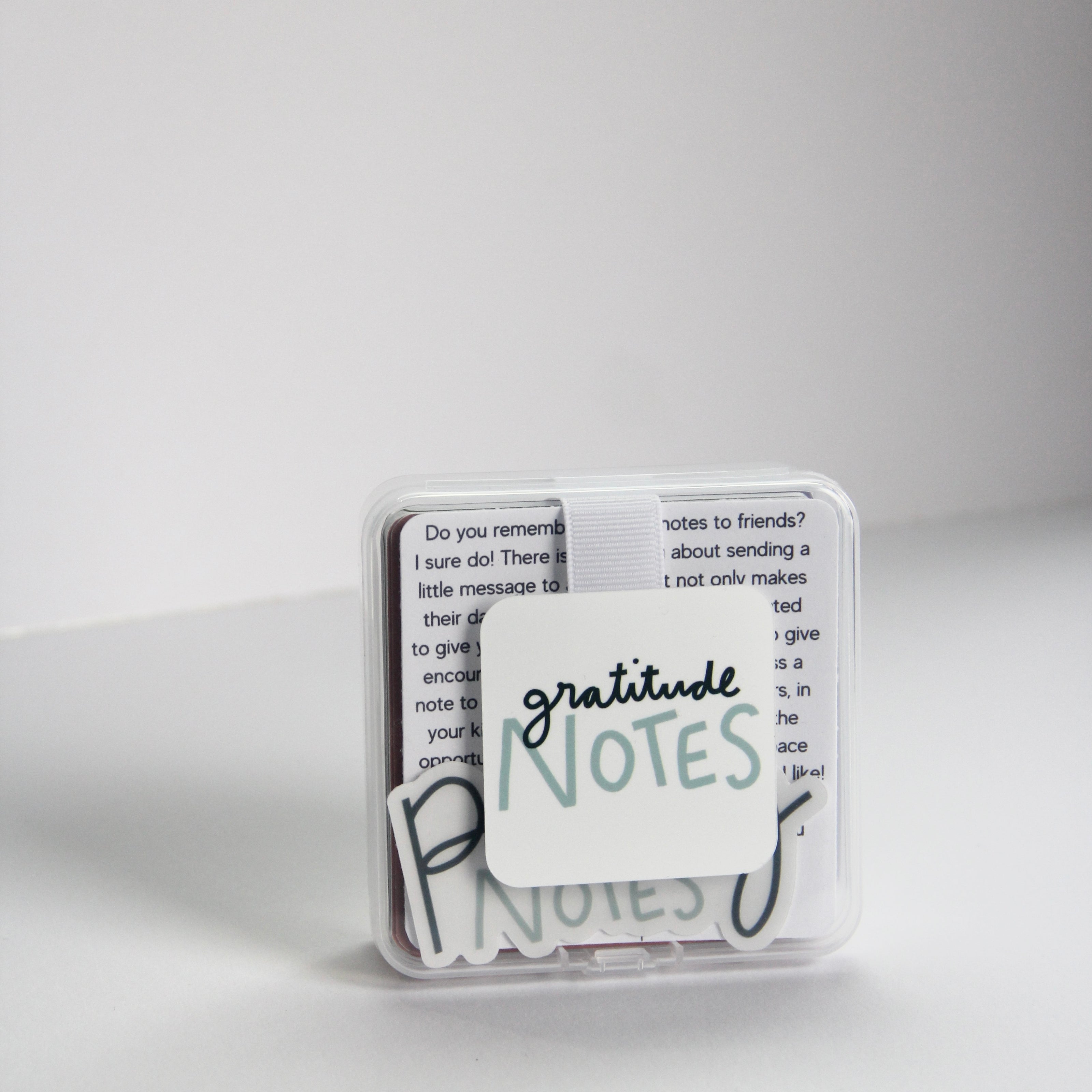 A small, clear box labeled "Gratitude Notes" contains note cards filled with gratitude affirmations. The background is plain white, and a few cards are slightly visible inside the box.