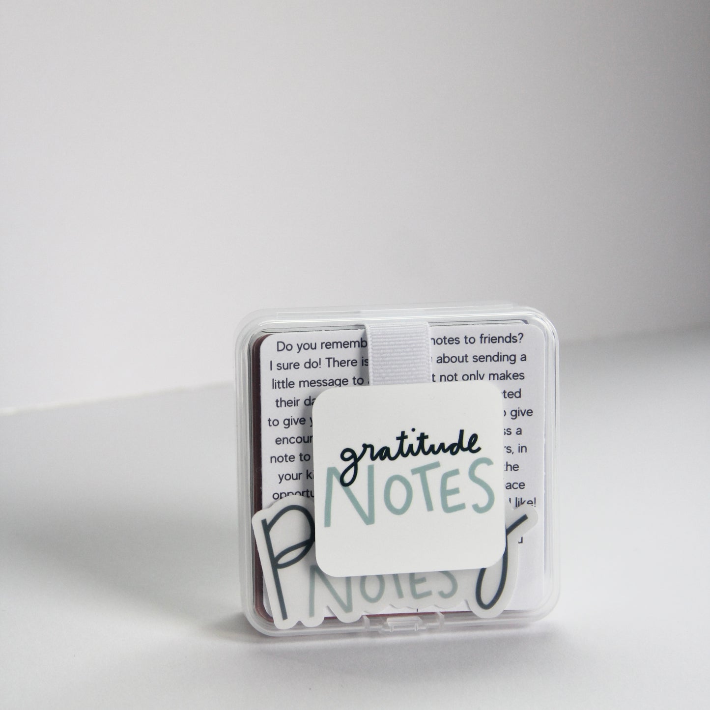 A small, clear box labeled "Gratitude Notes" contains note cards filled with gratitude affirmations. The background is plain white, and a few cards are slightly visible inside the box.