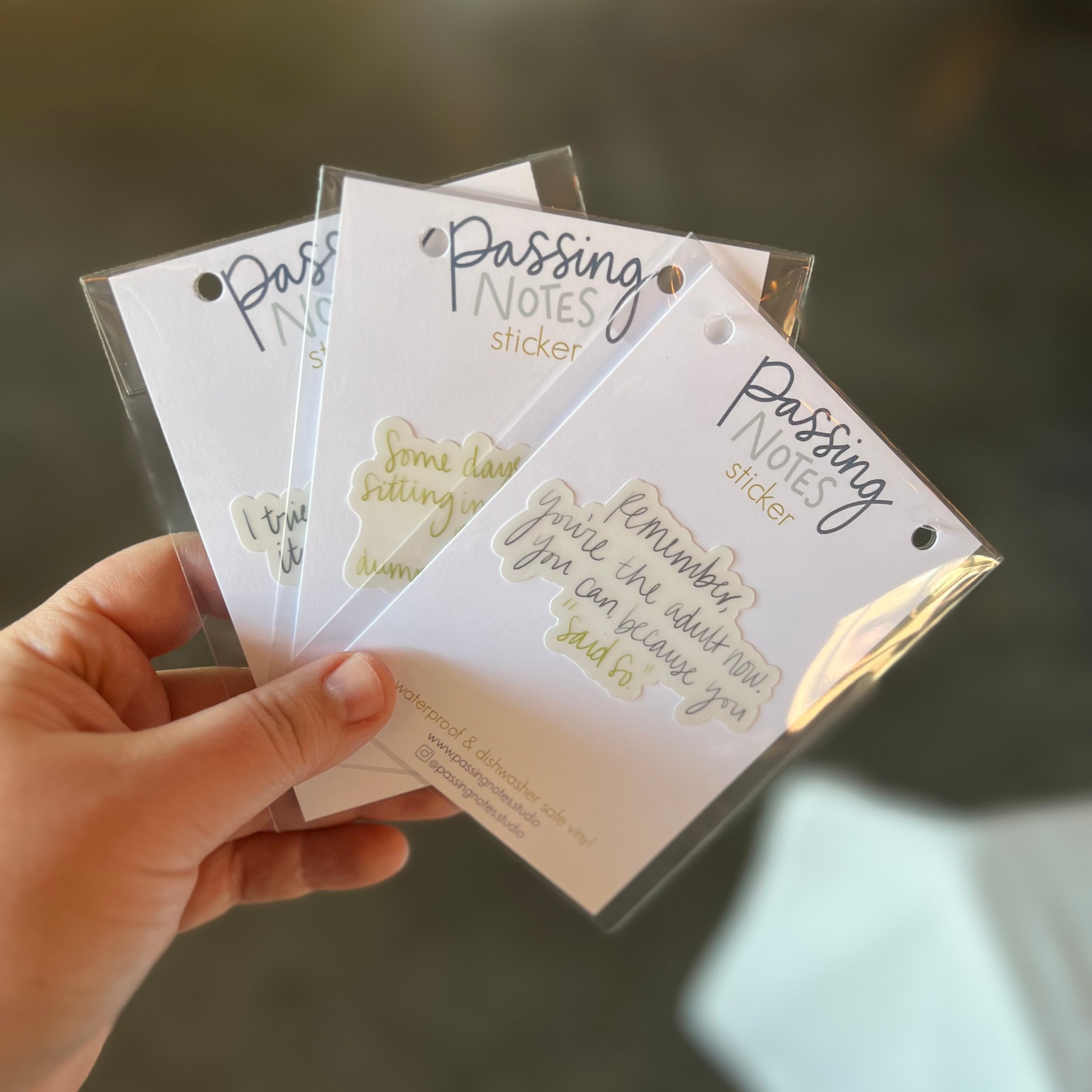 A hand holds three "Passing Notes" sticker packs, each featuring handwritten-style text in varying colors. These unique funny stickers stand out against the blurred, neutral background.