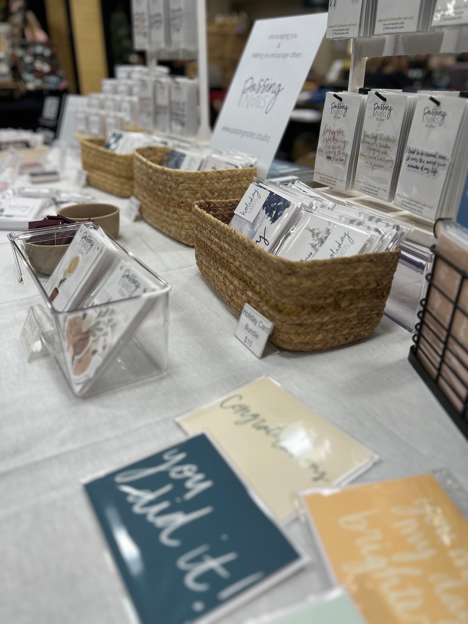 A table display at a market in Minnesota features baskets filled with greeting cards and note packs. Cards with messages like "You did it!" and "Congratulations" are visible. Signage for "Passing Notes" is in the background, with a cozy and inviting setup.