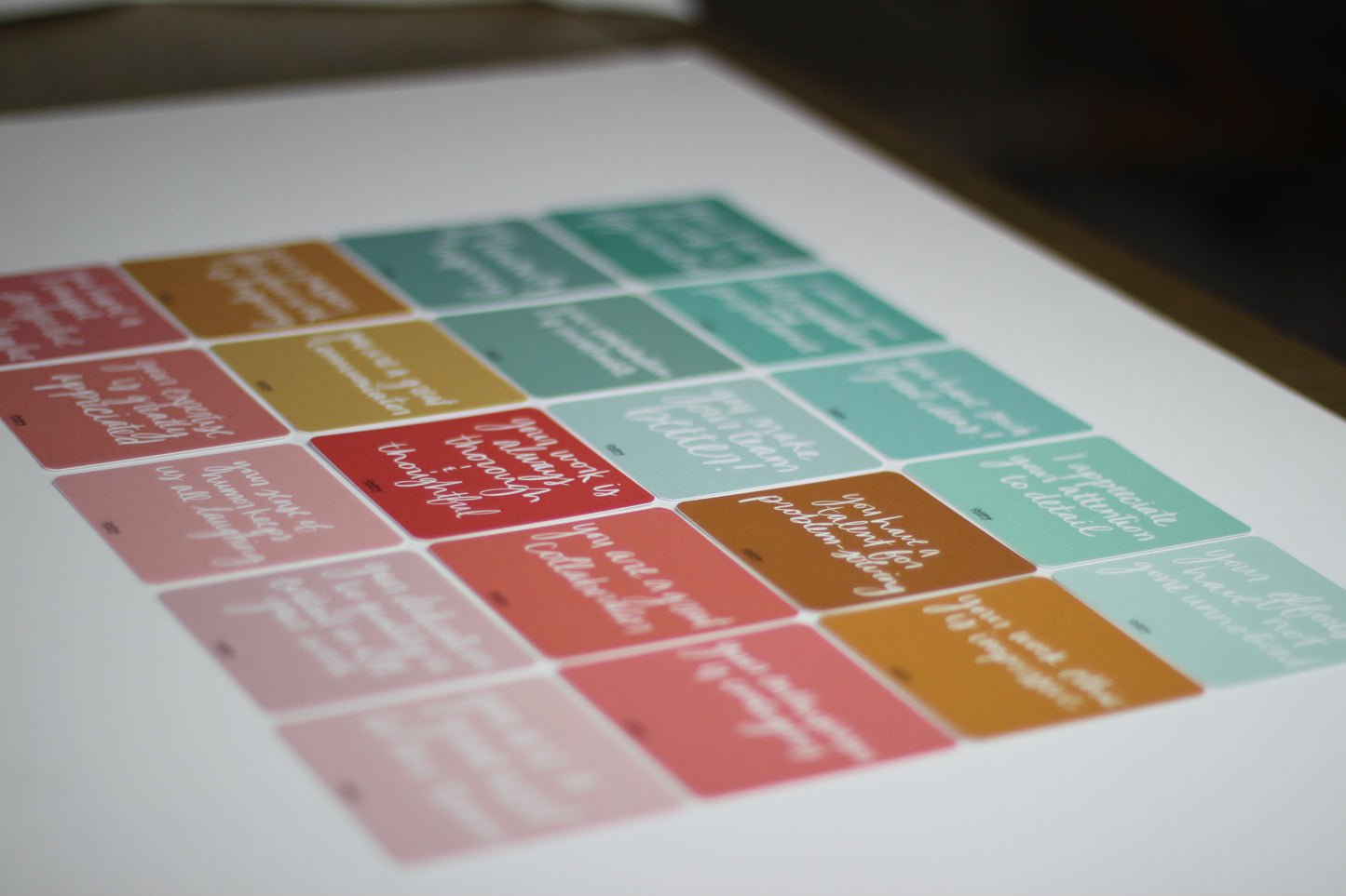 A sheet of colorful, rectangular stickers designed like note cards with motivational quotes is arranged on a white surface. The stickers come in shades of pink, green, orange, and brown, each featuring white text in various fonts—ideal for sharing encouragement and appreciation with coworkers.