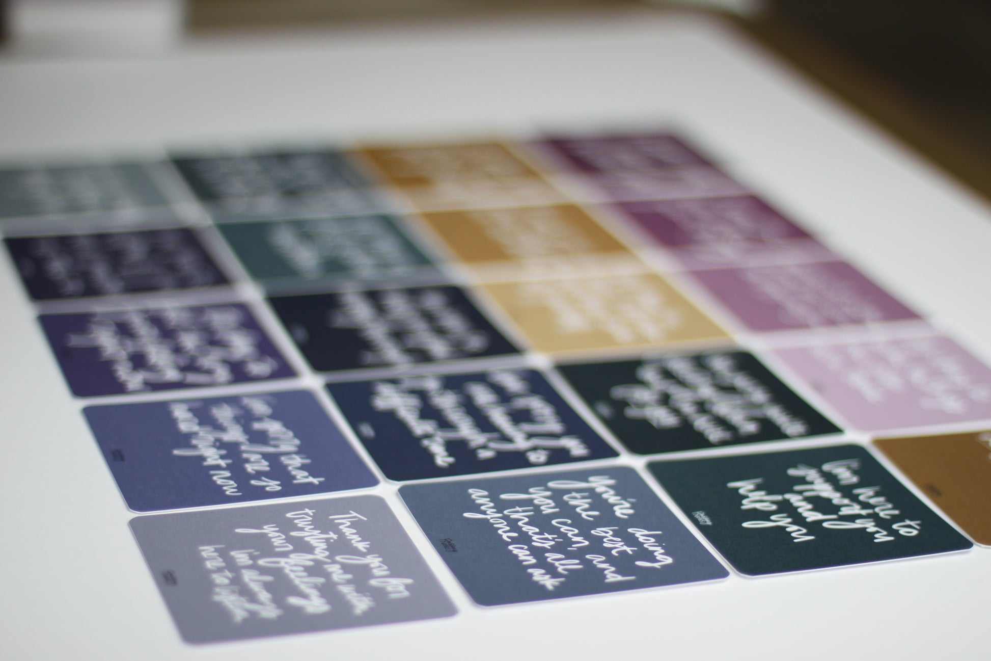 An array of colorful square empathy note cards are laid out on a light surface, each with different handwritten messages in white script. Featuring purples, blues, and greens, these encouraging notes blend warmth and care. The text remains mostly unreadable due to the focus.