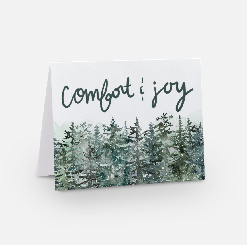 A holiday greeting card displaying the words "comfort & joy" in cursive font above a watercolor illustration of evergreen trees. This artful keepsake stands against a white background, perfect for adding a personalized message inside.