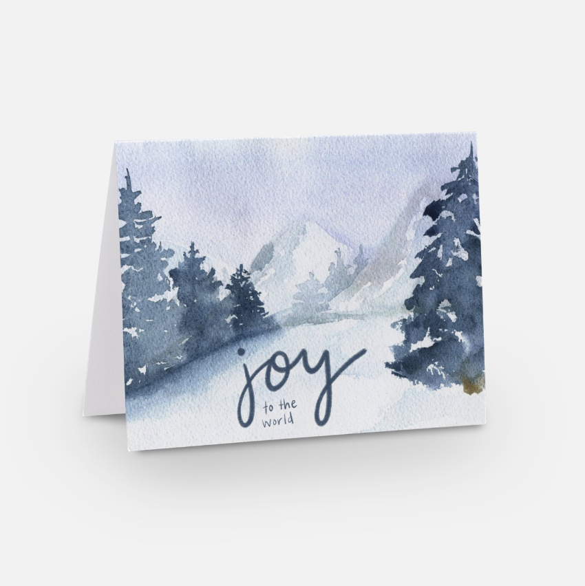 A holiday greeting card featuring festive artwork of a watercolor winter scene with snowy trees and mountains. "Joy to the World" is elegantly scripted across the landscape. This holiday card is folded and displayed on a plain background, perfect for adding personalized messages inside.