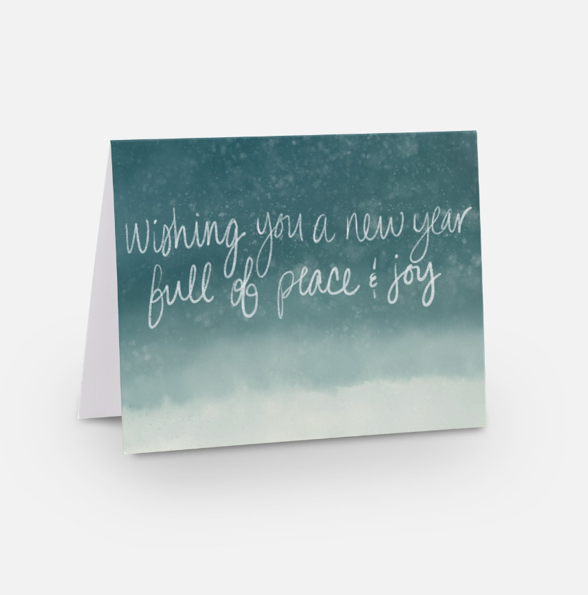 A New Year card against a soft blue gradient background features handwritten text: "Wishing you a new year full of peace & joy." The festive design conveys heartfelt wishes as the card stands slightly open on a plain surface.